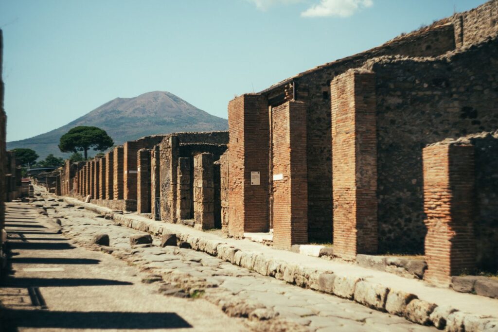 Ancient cobblestone street in Pompeii with preserved Roman buildings and Mount Vesuvius visible in the background.