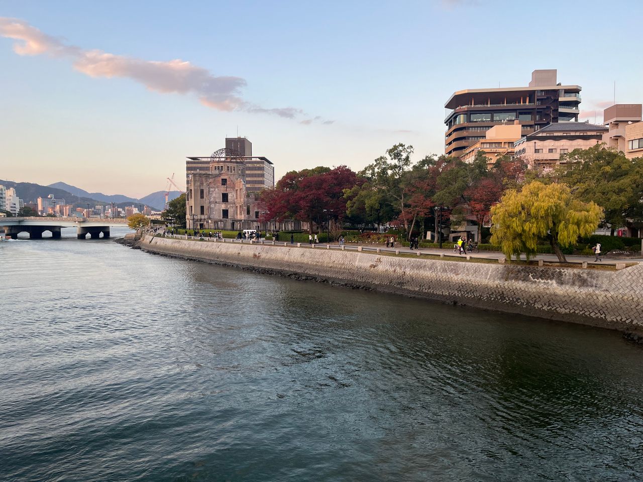 Atomic Bomb Dome beside the Motoyasu River in Hiroshima, Japan, during sunset with autumn-colored trees, waterfront promenade, and city buildings in the background.