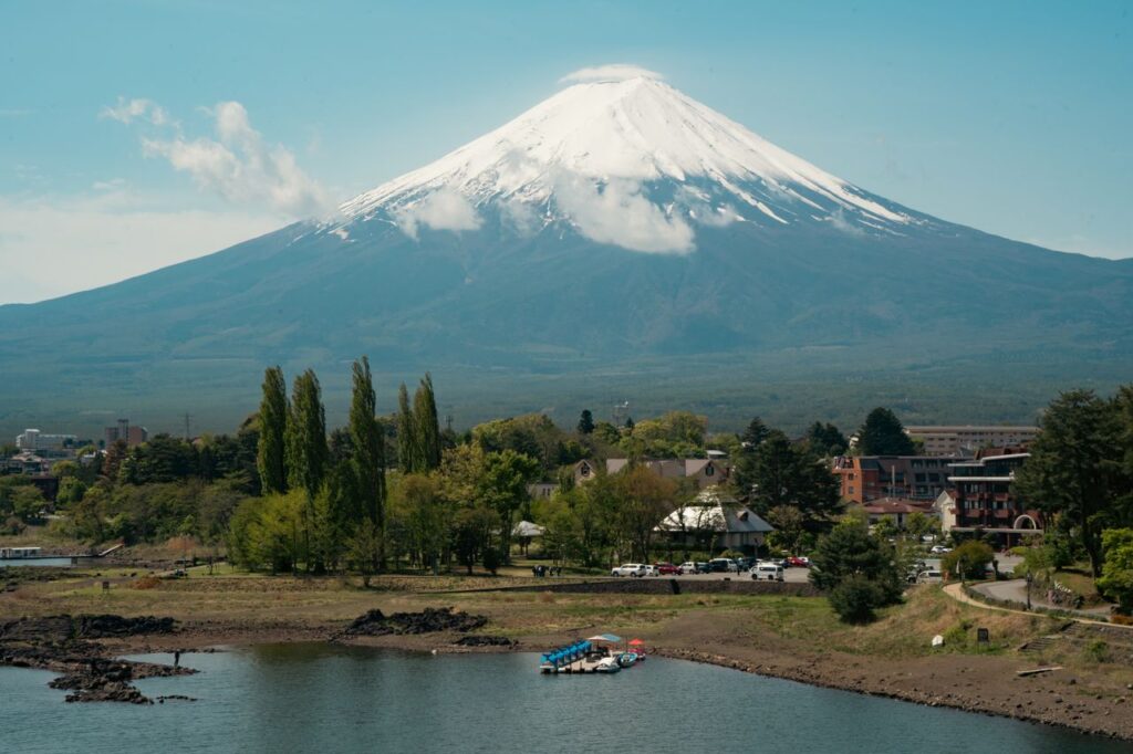 Panoramic view of snow-capped Mount Fuji towering above Lake Kawaguchiko, with lakeside trees, buildings, and calm waterfront scenery in Yamanashi Prefecture, Japan.