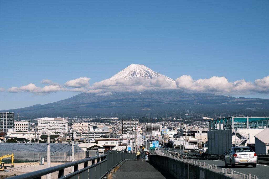 Snow-capped Mount Fuji towering above Fujinomiya City, with urban buildings, roads, and low clouds stretching across the mountain's lower slopes under a clear blue sky.
