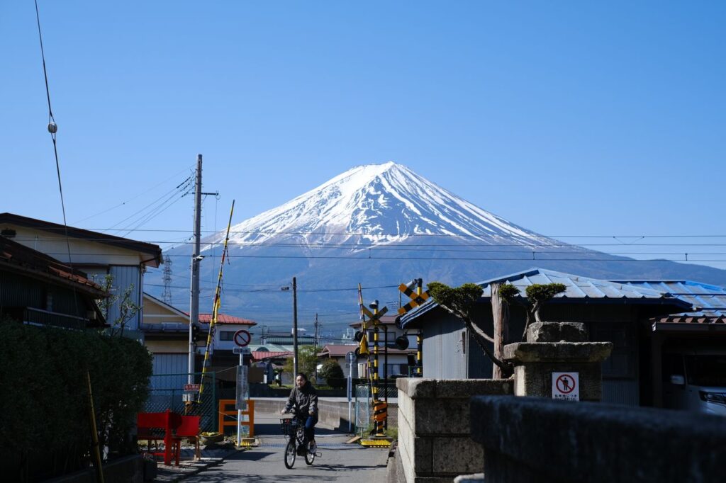 Mount Fuji covered in snow rising above a quiet residential street in Fujiyoshida, Japan, with a cyclist riding past traditional homes under a clear blue sky.