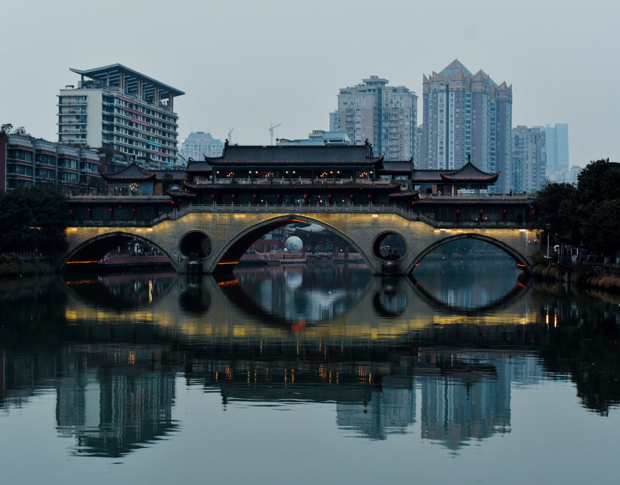 An illuminated traditional Chinese bridge spanning a calm river at dusk, with modern Chengdu skyline buildings reflected in the water behind it.