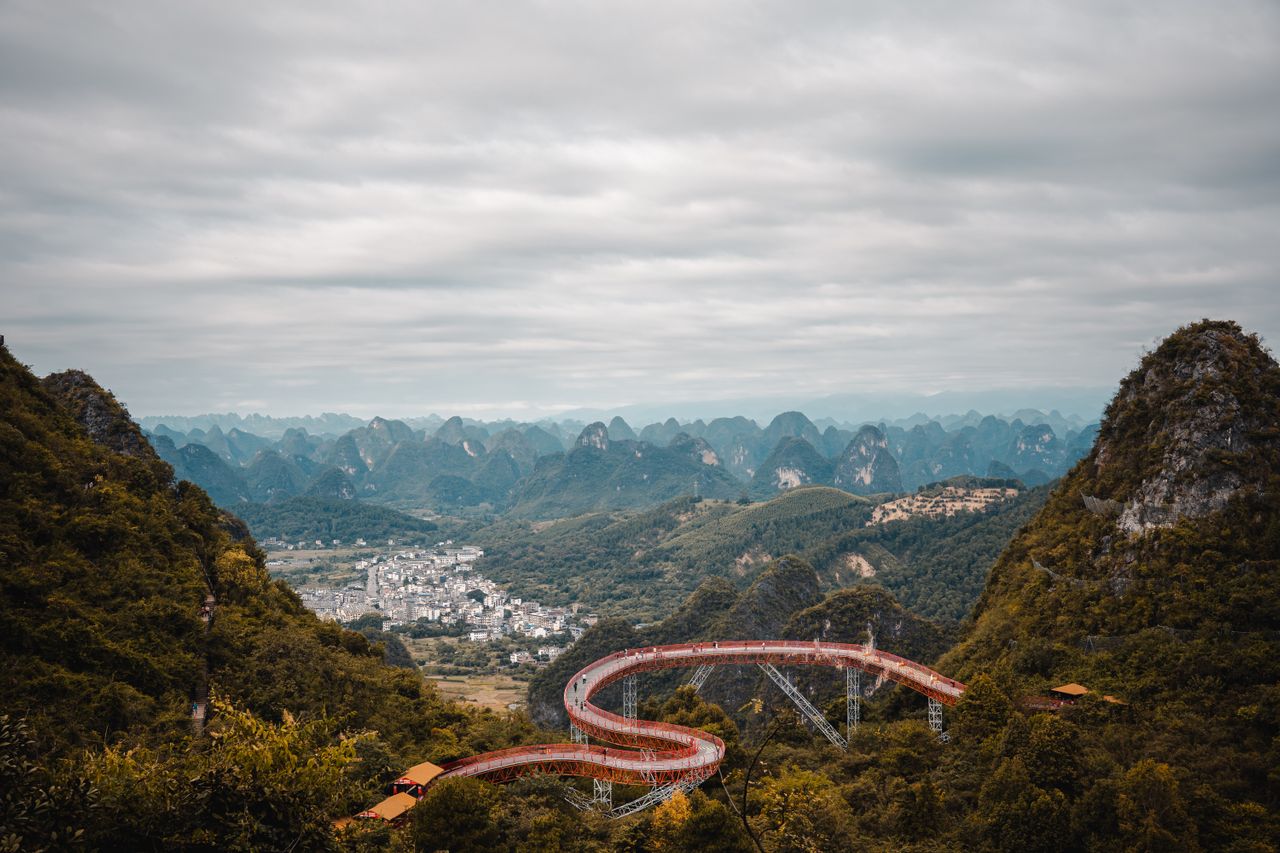 A panoramic view of a winding glass skywalk overlooking dramatic karst mountains and valleys in Guangxi, China, under a cloudy sky.