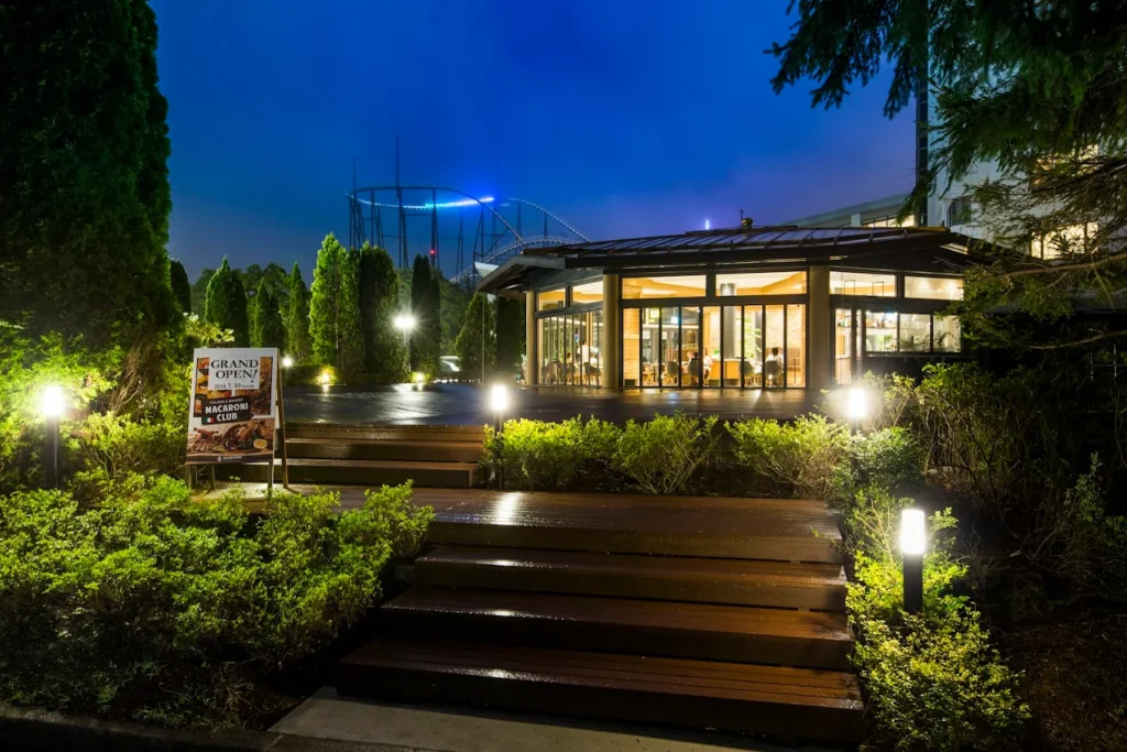Night view of Highland Resort Hotel & Spa in Fujiyoshida, Japan, with its warmly lit restaurant entrance, landscaped gardens, and Fuji-Q Highland roller coaster visible in the background.