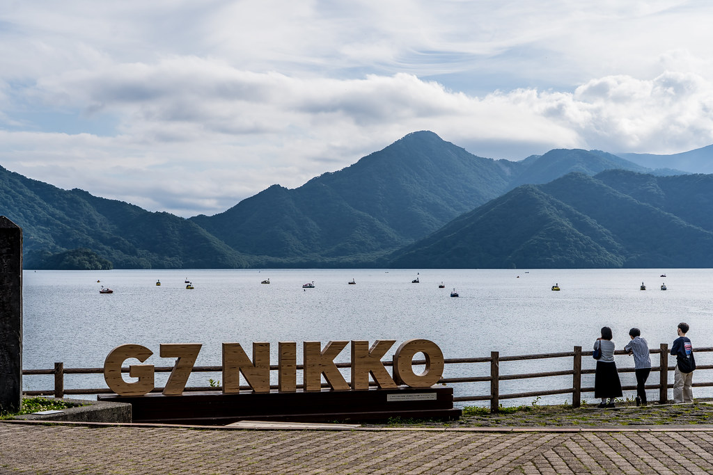 Lake Chuzenji in Nikko, Japan, with mountain views, calm waters, and the popular G7 Nikko sign along the lakeside promenade.