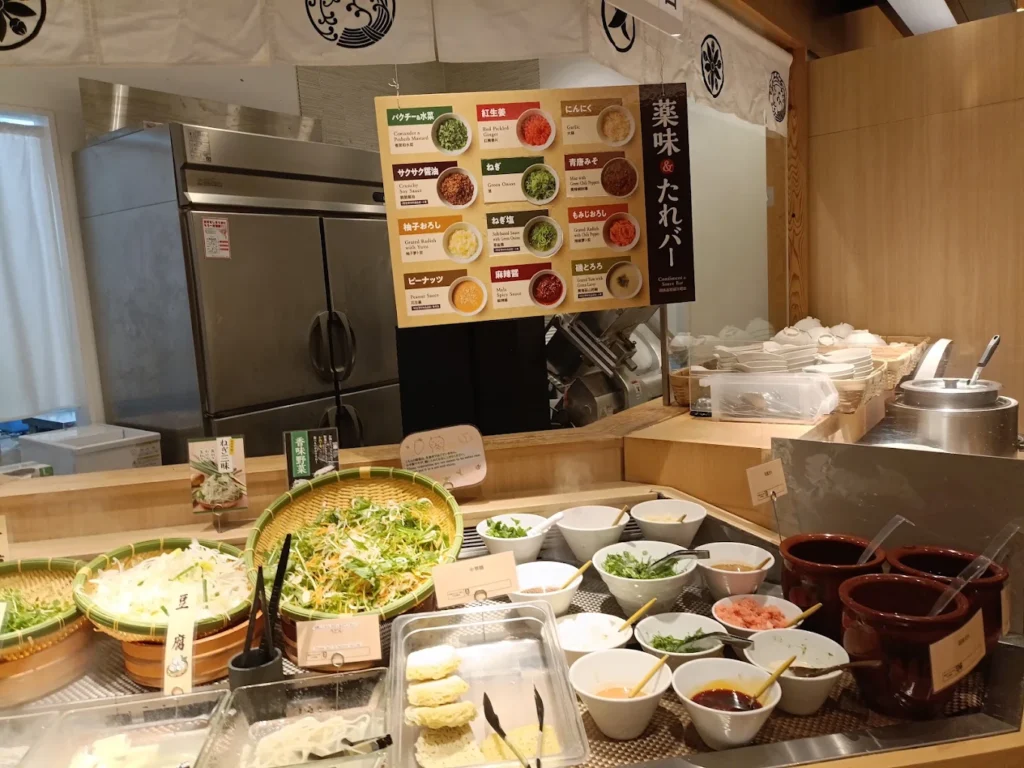 Fresh vegetable, sauce and condiment bar at Shabu Sai AEON Mall Kobe Kita, featuring unlimited vegetables and customizable dipping sauces for the all-you-can-eat shabu shabu buffet.
