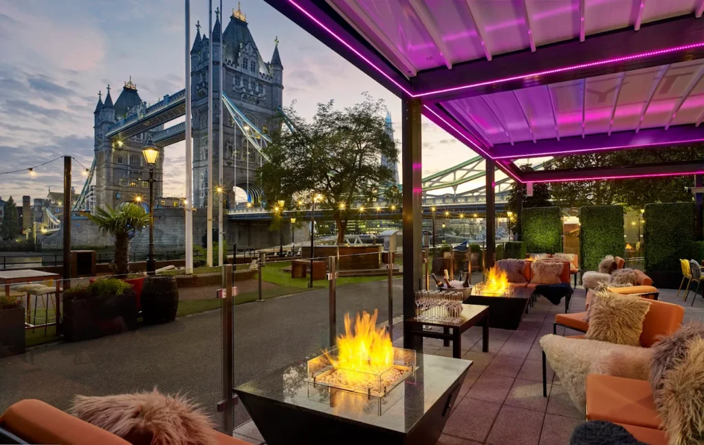 10 Best Muslim-Friendly Hotels (Halal) in London 10 Outdoor terrace at The Tower Hotel London overlooking the iconic Tower Bridge, featuring modern seating, fire pits, and views of the River Thames at dusk.