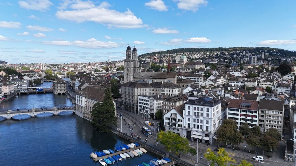 Panoramic view of Zurich, Switzerland, featuring the Limmat River, historic Old Town, Grossmünster Church, and the city skyline under a bright blue sky.