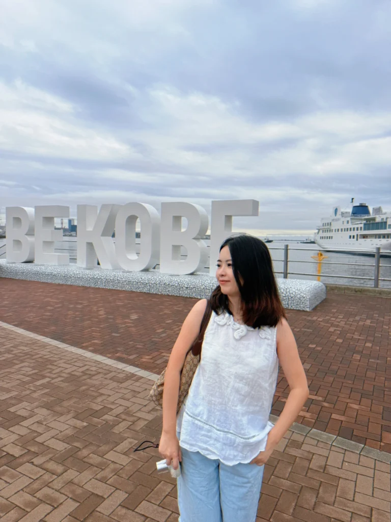 7-Day Osaka, Kyoto and Kobe Itinerary: Food, Shopping and Summer Sights 10 Woman posing beside the large BE KOBE sign at Meriken Park with Kobe Harbour and a sightseeing ship in the background.