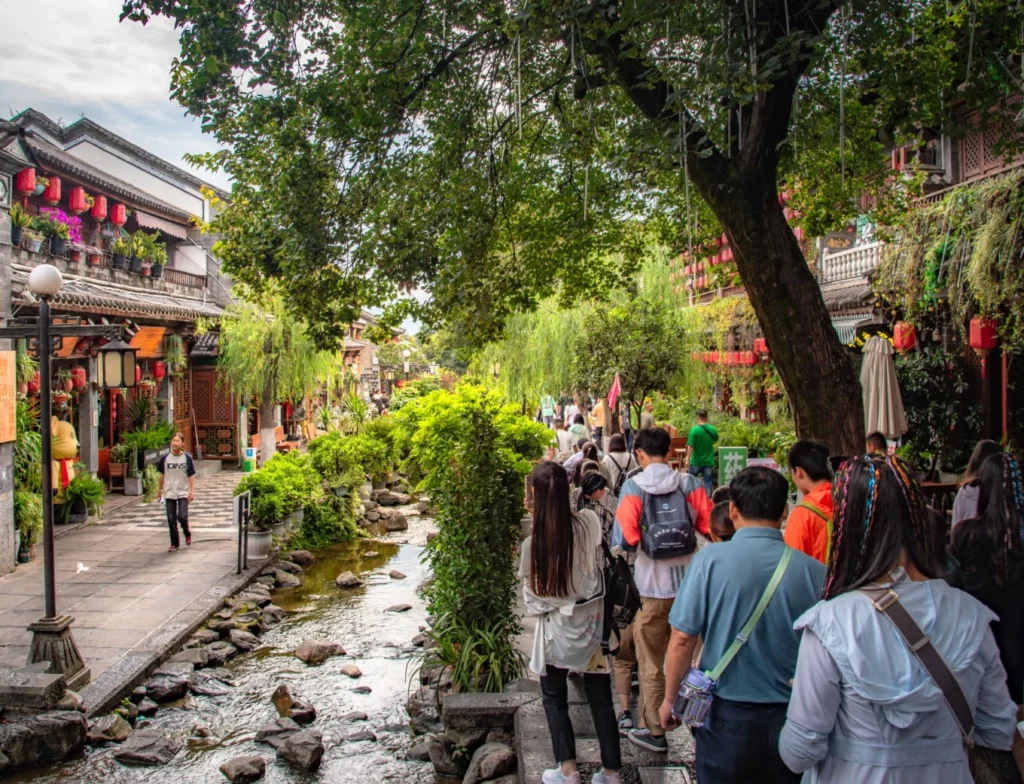 Where to Stay in Dali: 6 Best Areas & Hotels 6 Crowded pedestrian street beside a tree-lined stream in Dali Ancient Town, with traditional buildings, red lanterns and shops.
