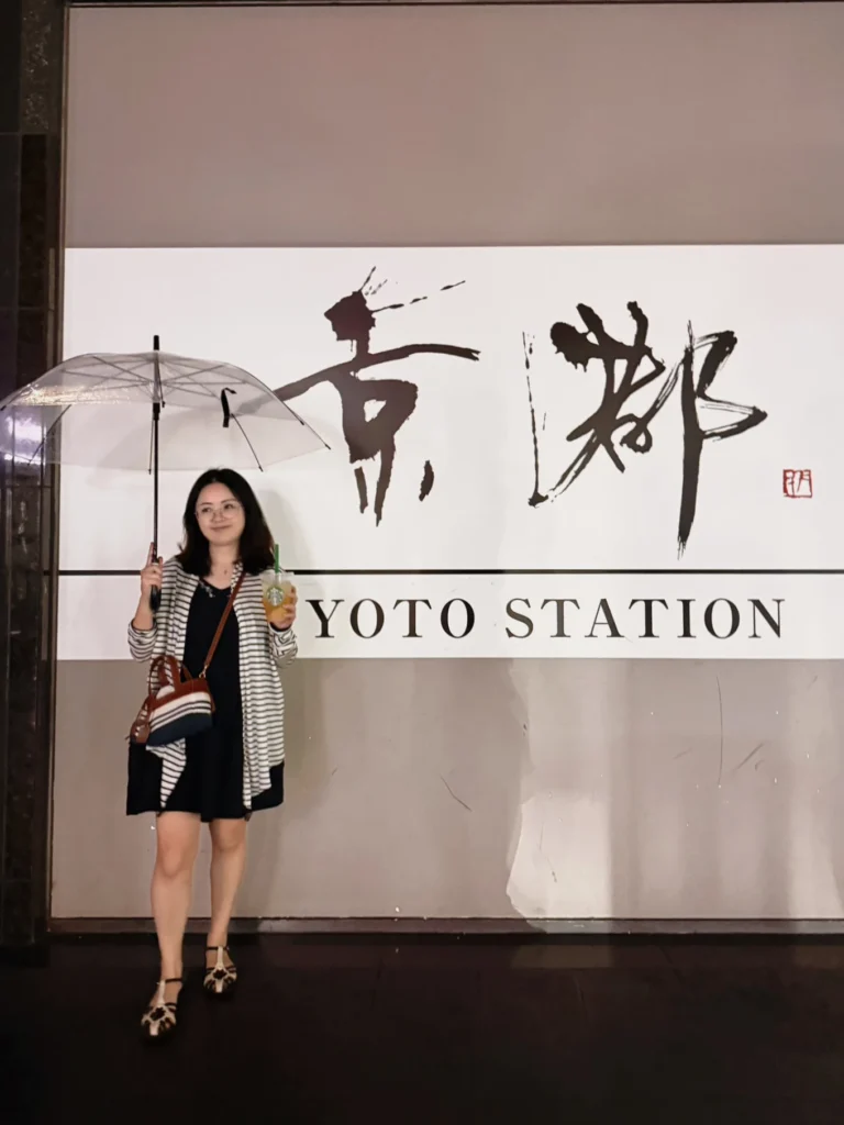 7-Day Osaka, Kyoto and Kobe Itinerary: Food, Shopping and Summer Sights 19 Woman holding an umbrella and drink in front of the Kyoto Station sign on a rainy evening.