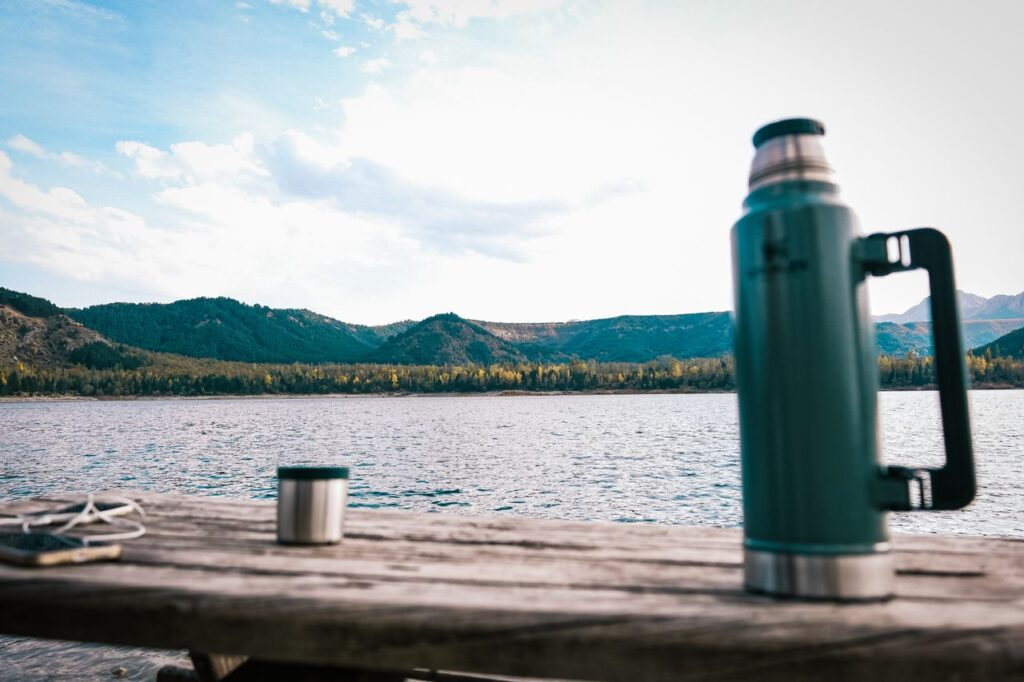 Green travel thermos flask and drinking cup on a wooden table beside a mountain lake.