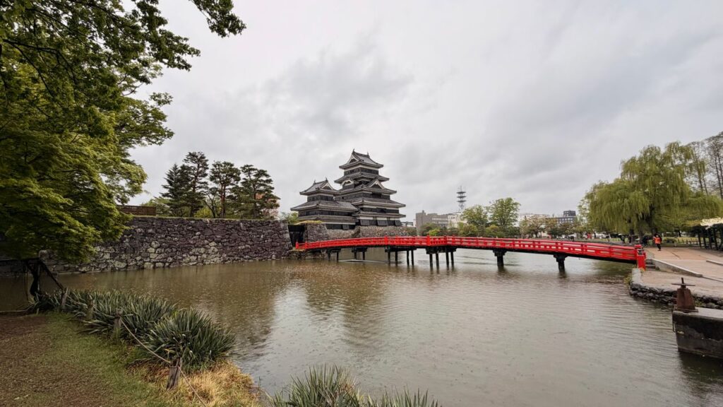 Matsumoto Castle viewed across its moat with the iconic red bridge, surrounded by lush greenery under a cloudy sky in Nagano, Japan.