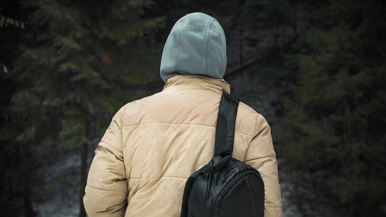 Traveler wearing a hooded jacket and backpack walking through light rain in a forest, illustrating the importance of a packable travel rain jacket.