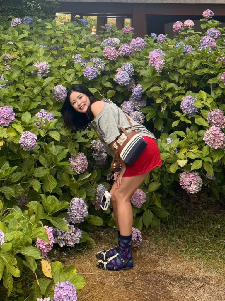 A woman with dark hair leans forward smiling in front of a lush, dense bush of purple and pink hydrangeas. She wears an off-the-shoulder grey shirt, a short red skirt, patterned purple socks, and black strappy hybrid ballet flats. A small striped bag with brown handles hangs over her shoulder, and she holds a compact white camera.