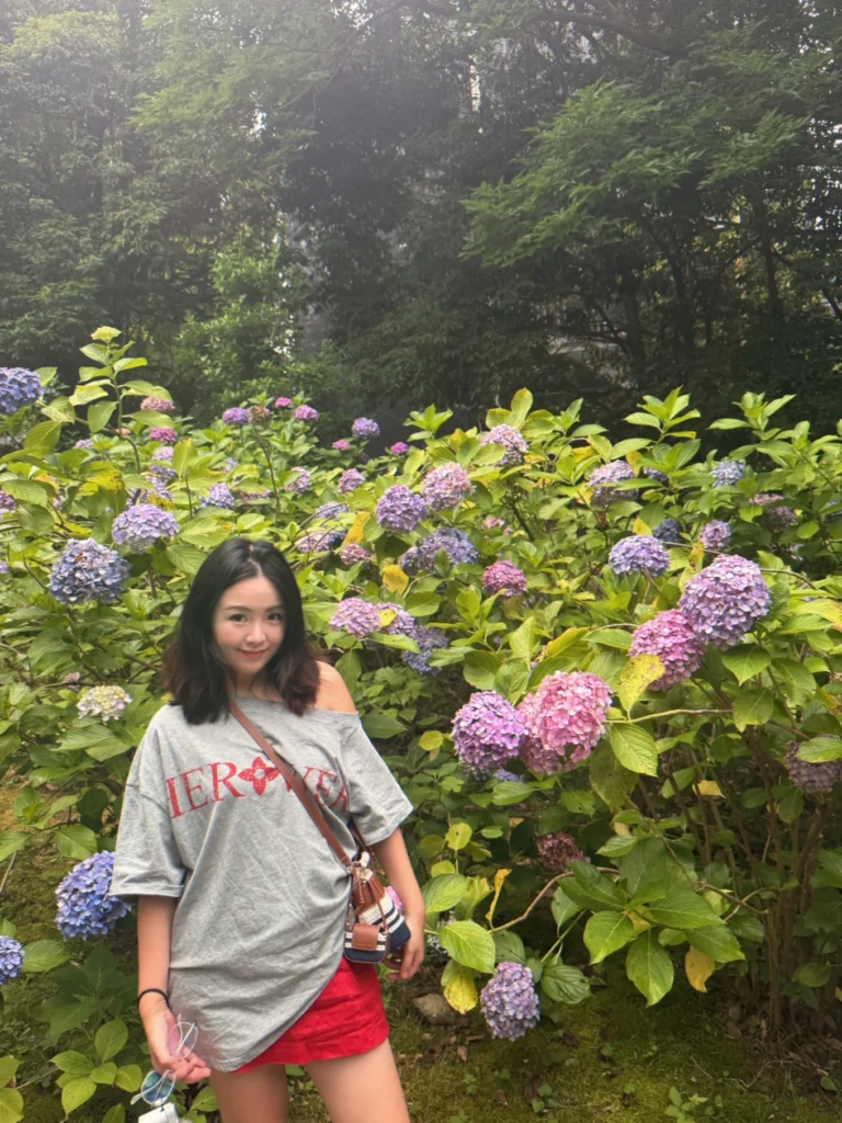 A woman with medium-length dark hair stands in front of a blooming hydrangea bush filled with purple and pink flowers. She wears an oversized, off-the-shoulder grey graphic T-shirt with red lettering, paired with a short red skirt. A small striped bag with a brown strap is worn crossbody style.